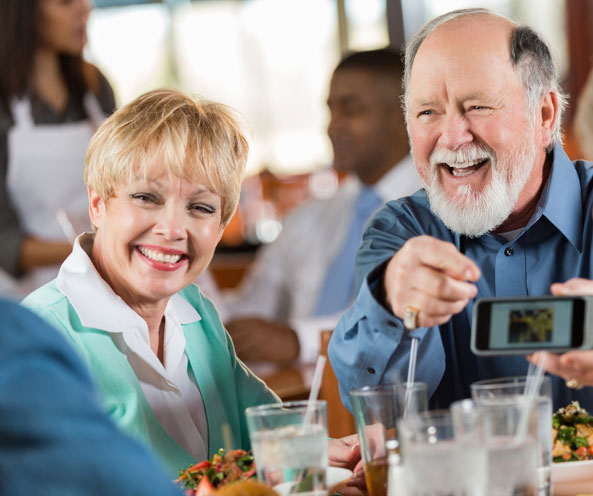 Older couple in community dining room sharing phone pictures and laughing.